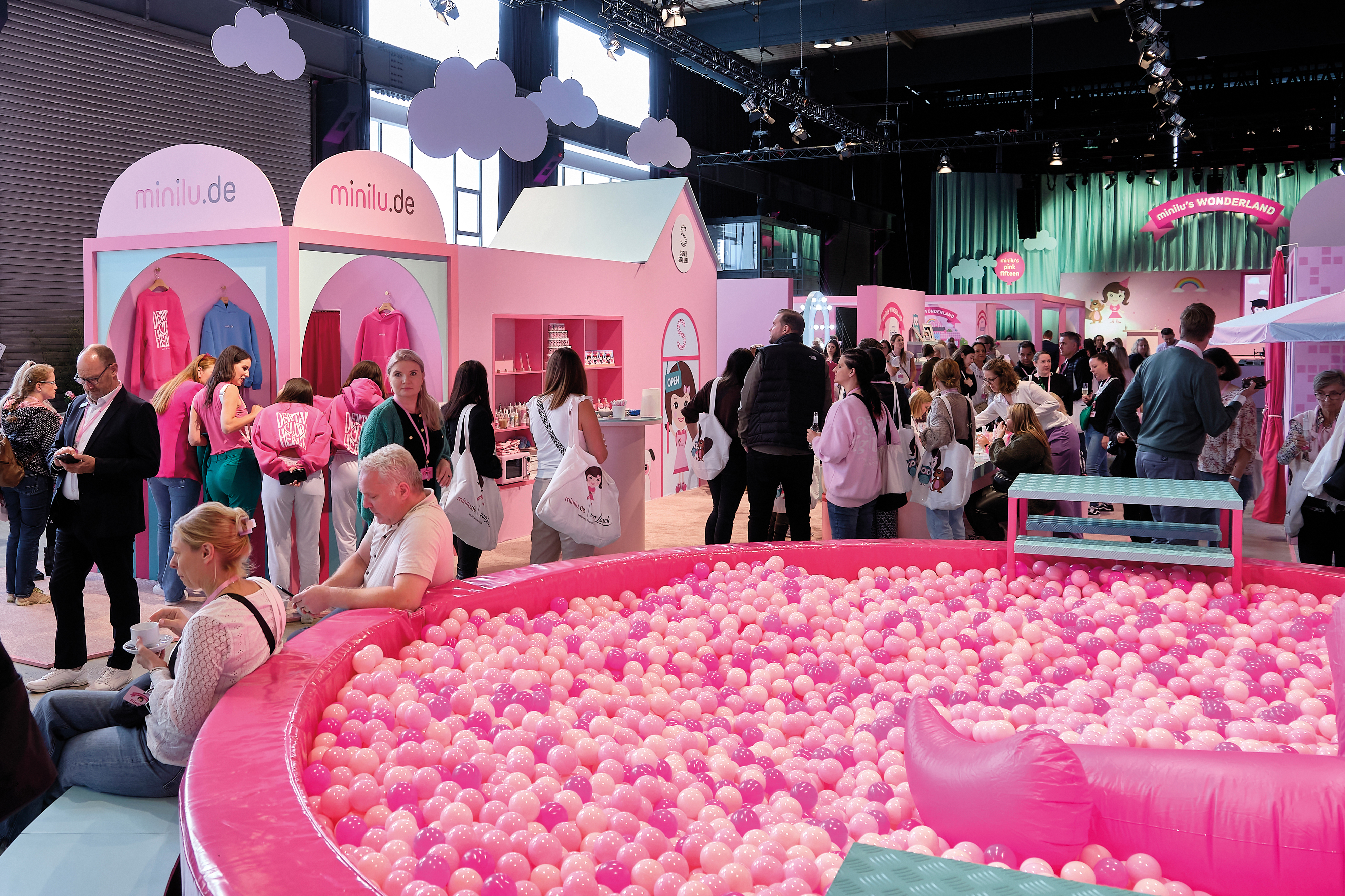  Die Szene ist eine lebendige Messehalle, beleuchtet von Scheinwerfern, mit hohen Decken und einem Betonboden. Im Vordergrund befindet sich ein riesiges, rundes Becken, das mit Tausenden von rosafarbenen und weißen Bällen gefüllt ist, und einige Personen sitzen auf dem Rand. Im Hintergrund ist eine große, rosafarbene Kulisse im Stil von kleinen, stilisierten Häusern zu sehen, mit Bögen und Schilder, die "minilu.de" zeigen. Viele Menschen bewegen sich durch den Eventbereich, und über ihnen schweben dekorative Wolken-Elemente.