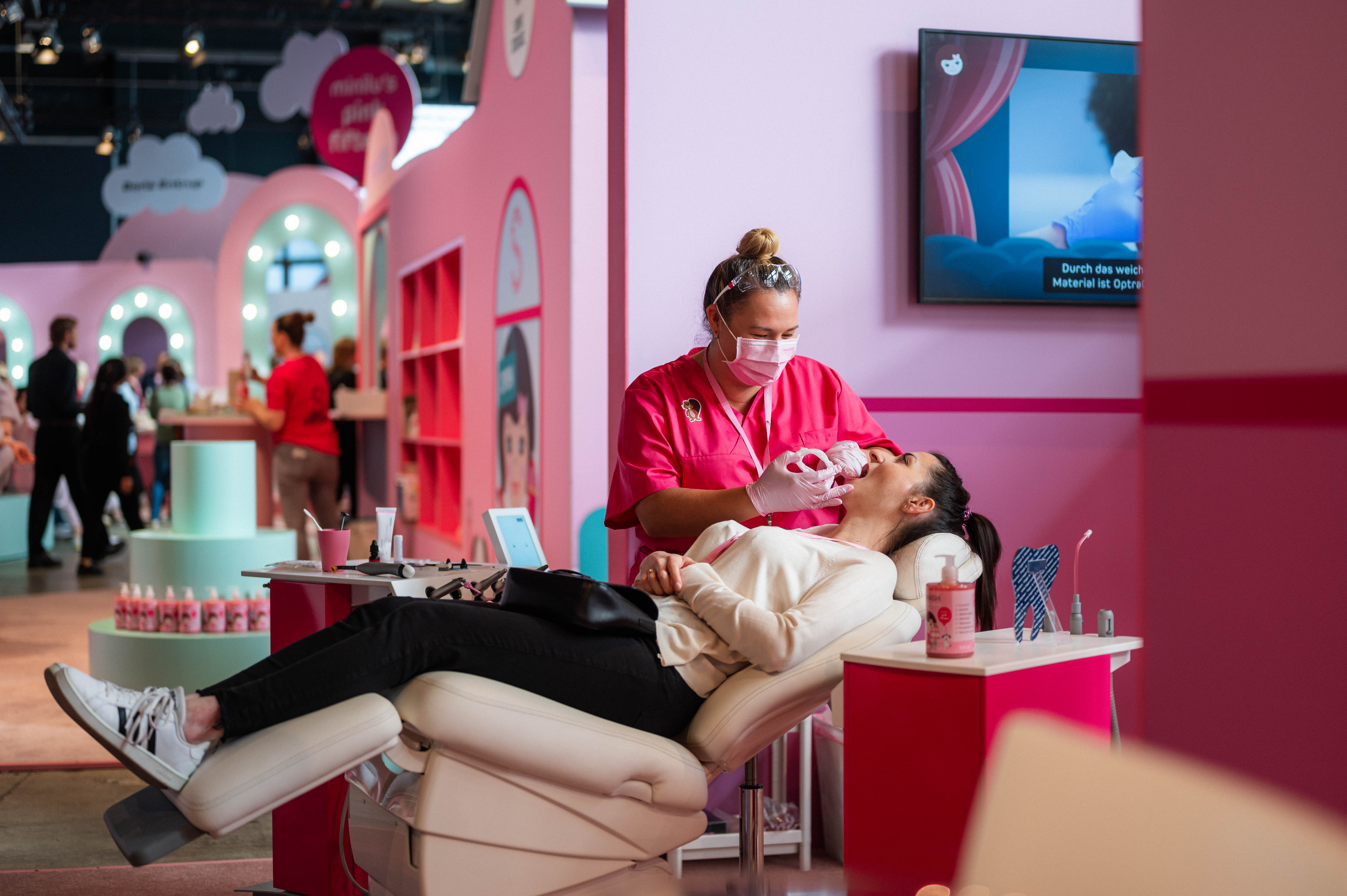 Eine Frau mit Maske und pinker Arbeitskleidung beugt sich über eine Person, die in einem Behandlungsstuhl liegt. Im Hintergrund ist ein großer Bildschirm zu sehen, der eine Produktpräsentation zeigt. Die Szene ist Teil des Events, das in rosafarbenen und weißen Tönen gehalten ist.