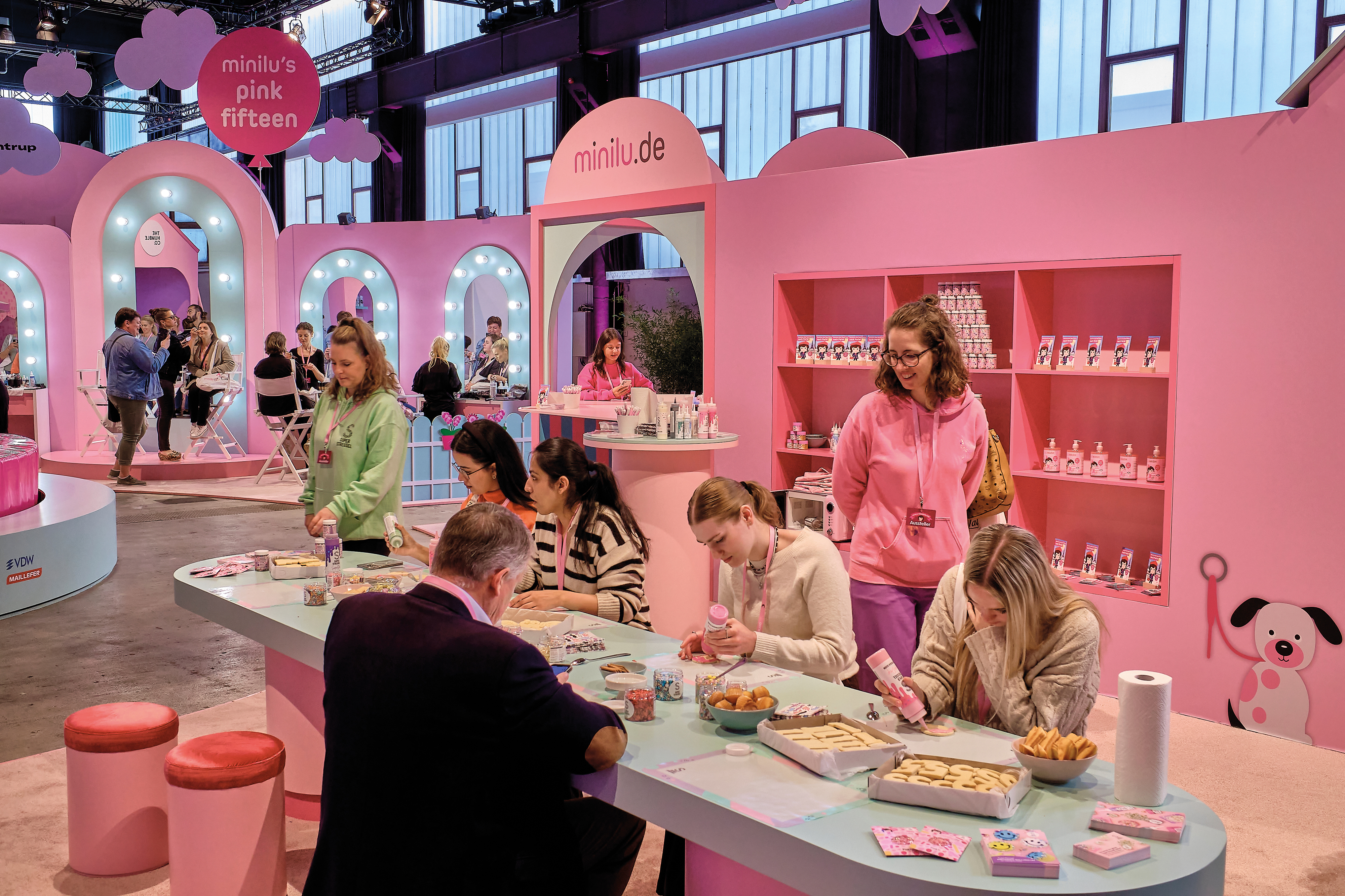 Mehrere Personen sitzen an einem hellblauen Tisch in der Eventkulisse "minilu's Wonderland" und verzieren Kekse. Eine Person im Vordergrund trägt ein hellgrünes Sweatshirt, eine andere eine rosa Kapuzenjacke. Im Hintergrund befindet sich ein pinkfarbener Stand mit Regalen voller Produkte und dem Schriftzug "minilu.de".