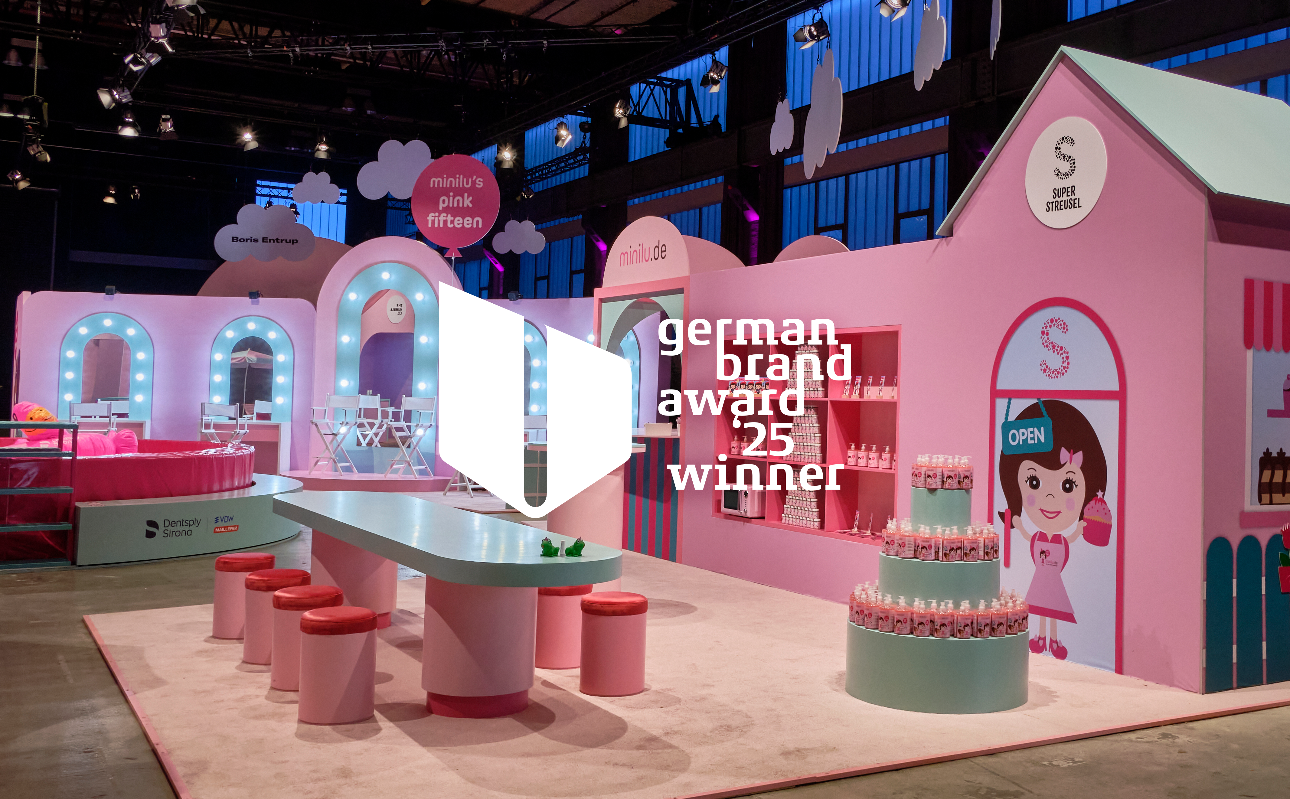 Eine Innenansicht des minilu's Wonderland Events. Im Vordergrund steht eine rosafarbene Eventkulisse im Stil eines Spielhauses oder eines Ladens mit dem Logo "Super Streusel". Es gibt eine Bar mit Hockern, einen Schminkbereich mit beleuchteten Spiegeln und eine Torten-Installation aus Produktflaschen. Über der Szene schweben rosafarbene und weiße Wolken-Elemente. Auf dem Bild ist ein transparentes Overlay mit der Aufschrift "german brand award '25 winner"
