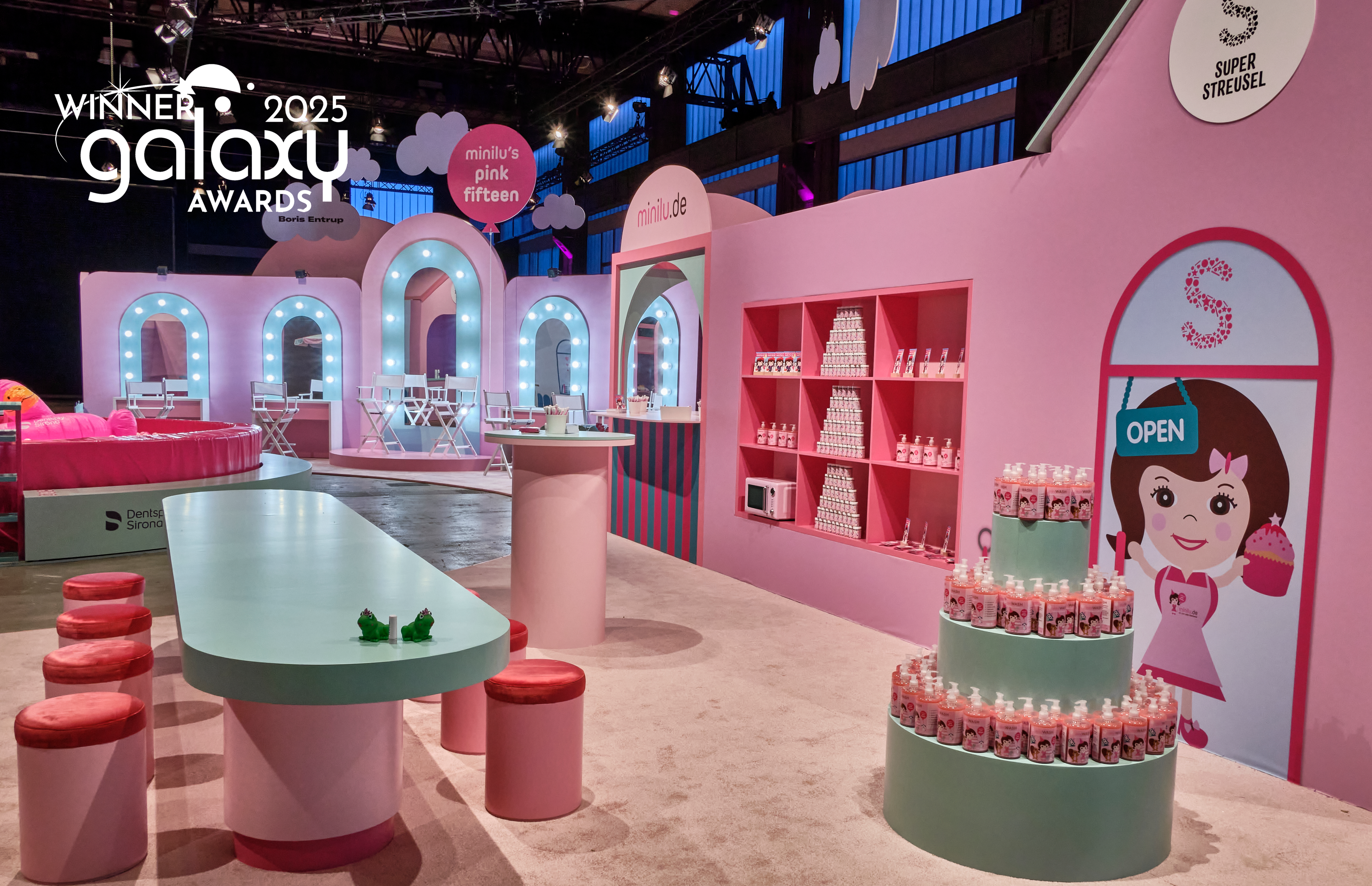 Eine Innenansicht des minilu's Wonderland Events. Im Vordergrund steht eine rosafarbene Eventkulisse im Stil eines Spielhauses oder eines Ladens mit dem Logo "Super Streusel". Es gibt eine Bar mit Hockern, einen Schminkbereich mit beleuchteten Spiegeln und eine Torten-Installation aus Produktflaschen. Über der Szene schweben rosafarbene und weiße Wolken-Elemente. Auf dem Bild ist ein transparentes Overlay mit der Aufschrift "german brand award '25 winner"