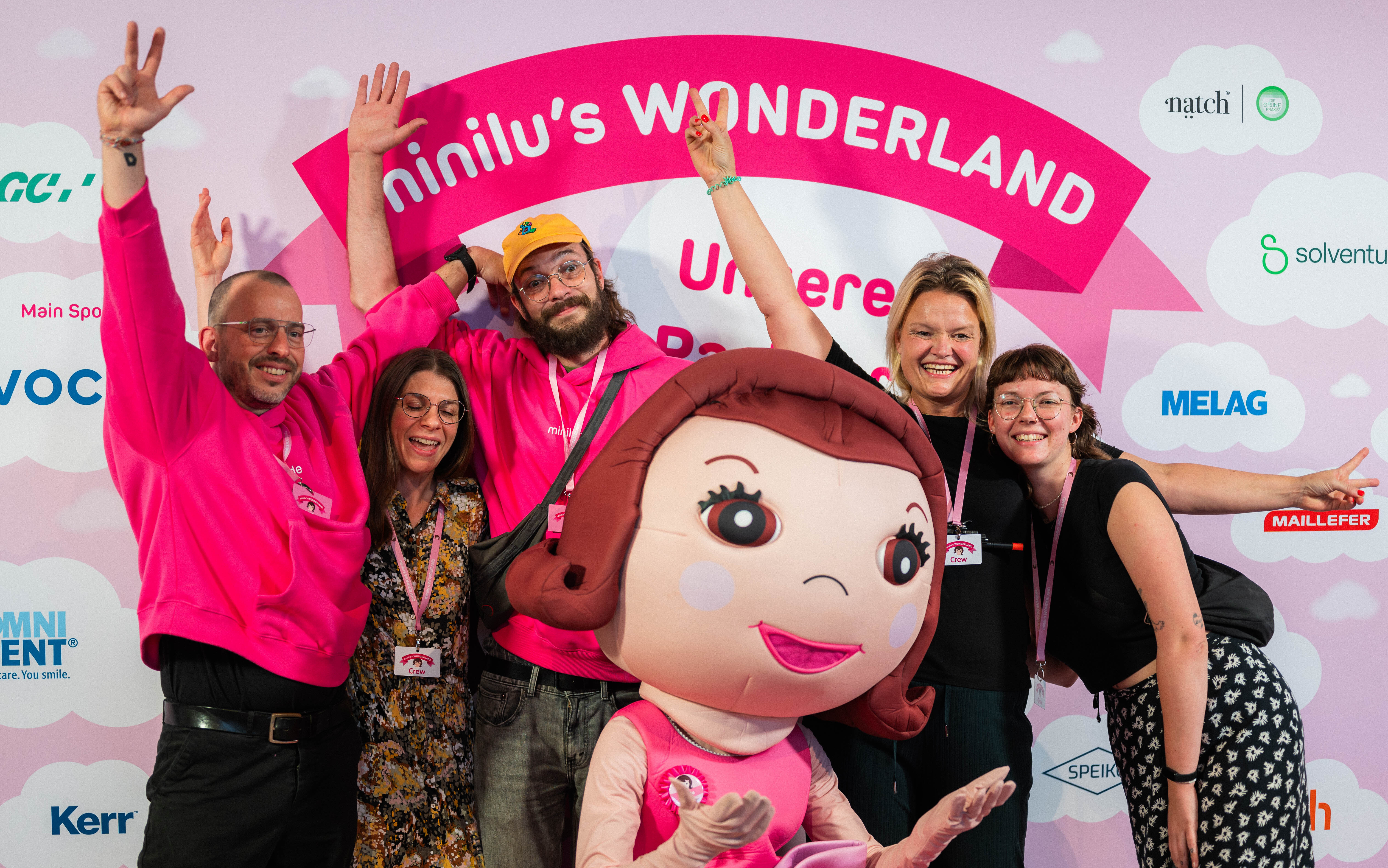 Eine Gruppe von sechs Personen in teils leuchtend pinker Kleidung posiert fröhlich mit der lebensgroßen Maskottchenfigur von miniluvor einer pinken Fotowand mit der Aufschrift "minilu's WONDERLAND". Die Personen gestikulieren mit den Armen und lächeln. Im Hintergrund sind Sponsorenlogos.